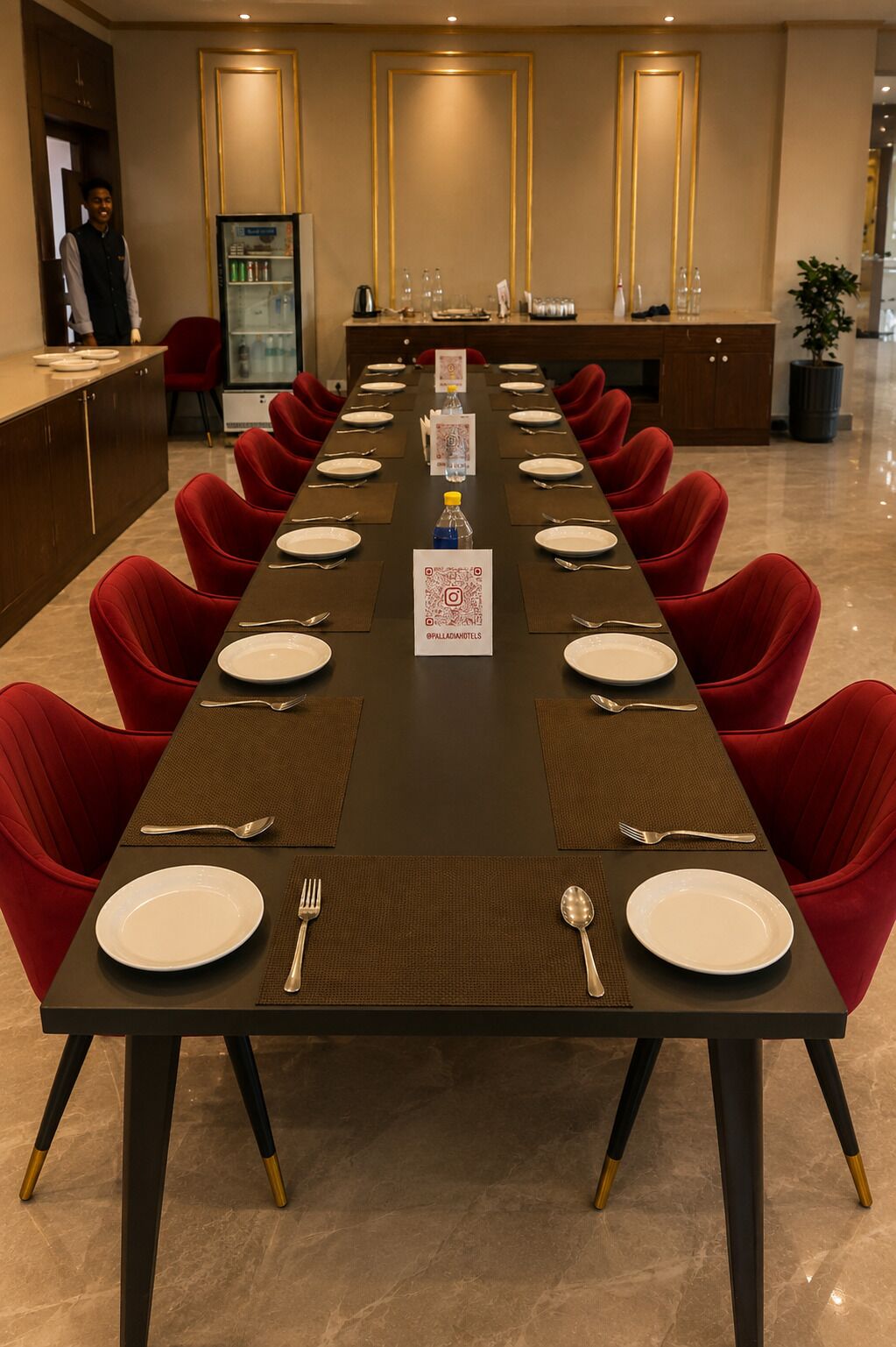 A conference room with a long table set for a meeting, red chairs, a minibar, and a reception area.