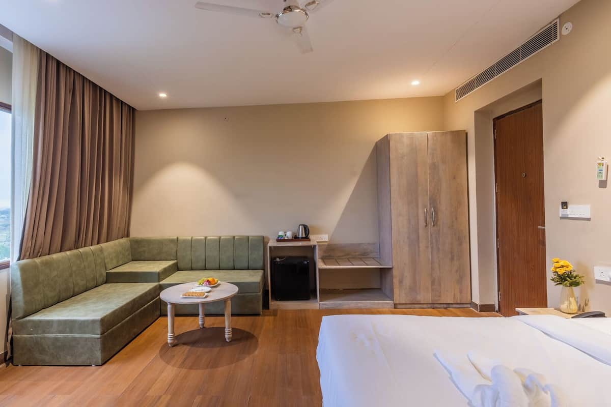 A hotel room with a green sofa, a small round table, a bed, a wardrobe, and a view of the outdoors.