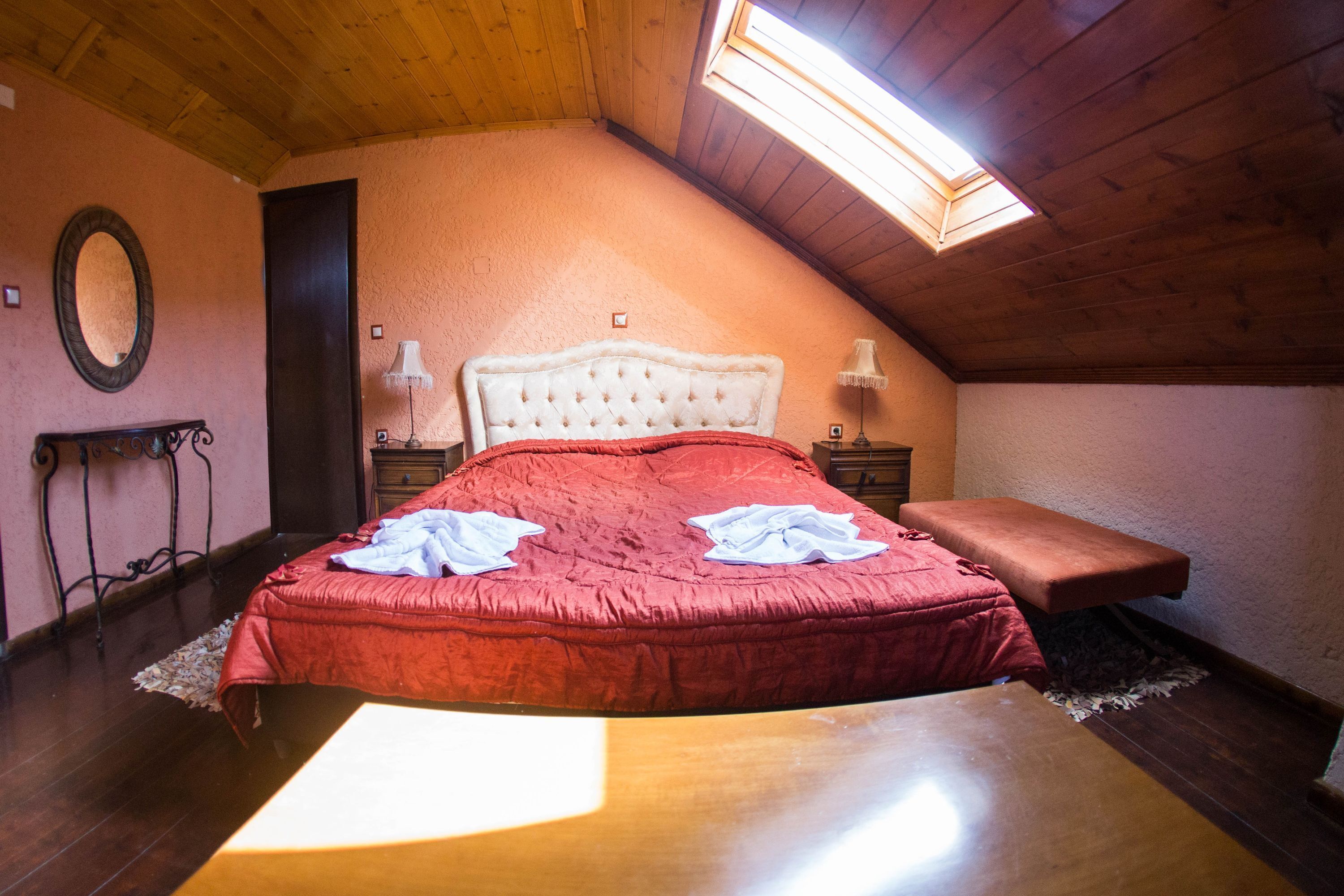 Een slaapkamer met een houten plafond, een schuine kap, een bed met een rode dekbedovertrek, twee bedlampen en een nachtkastje.