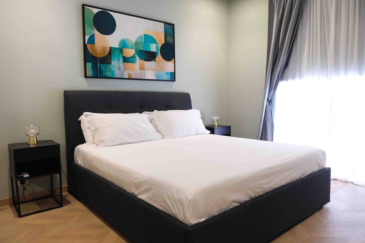 A bedroom with a large bed, a nightstand with a lamp, a wall art piece, and a window with curtains.