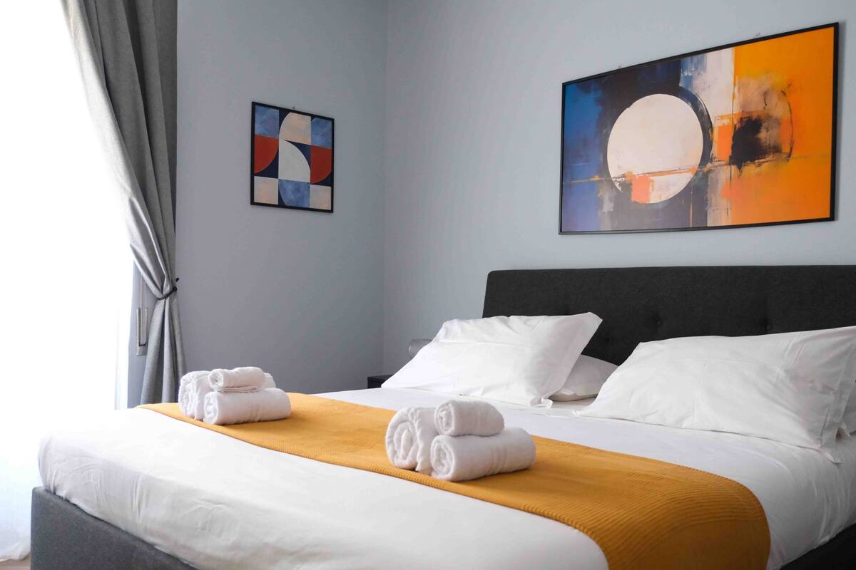 A neatly made bed with white linens and a mustard-colored throw, a gray headboard, and two abstract paintings on the wall.