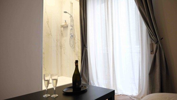 A hotel room with a bed, a table with two glasses and a bottle, a shower area, and a window with curtains. - Mazzini Suites Palermo (Palermo)
