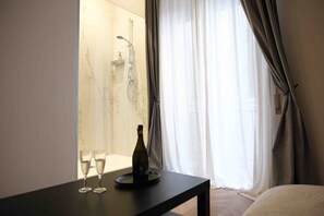 A hotel room with a bed, a table with two glasses and a bottle, a shower area, and a window with curtains. - Mazzini Suites Palermo (Palermo)