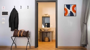 A hotel room with a wooden floor, a coat rack with a jacket, a Louis Vuitton bag on a small table, a bathroom with a sink and mirror, and a modern abstract painting on the wall. - Mazzini Suites Palermo (Palermo)