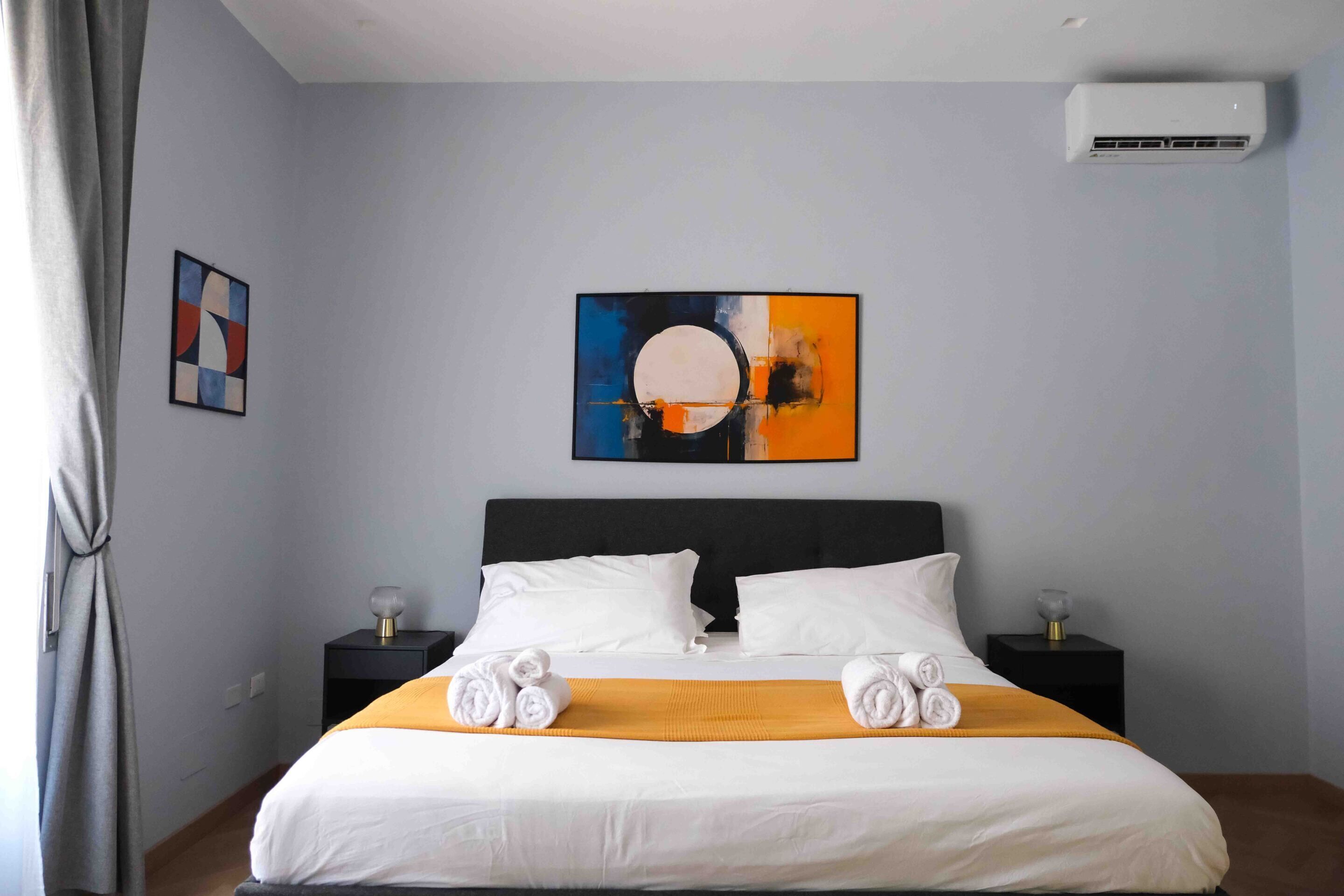 A hotel room with a large bed, two bedside tables, a wall-mounted air conditioner, and abstract art on the wall.