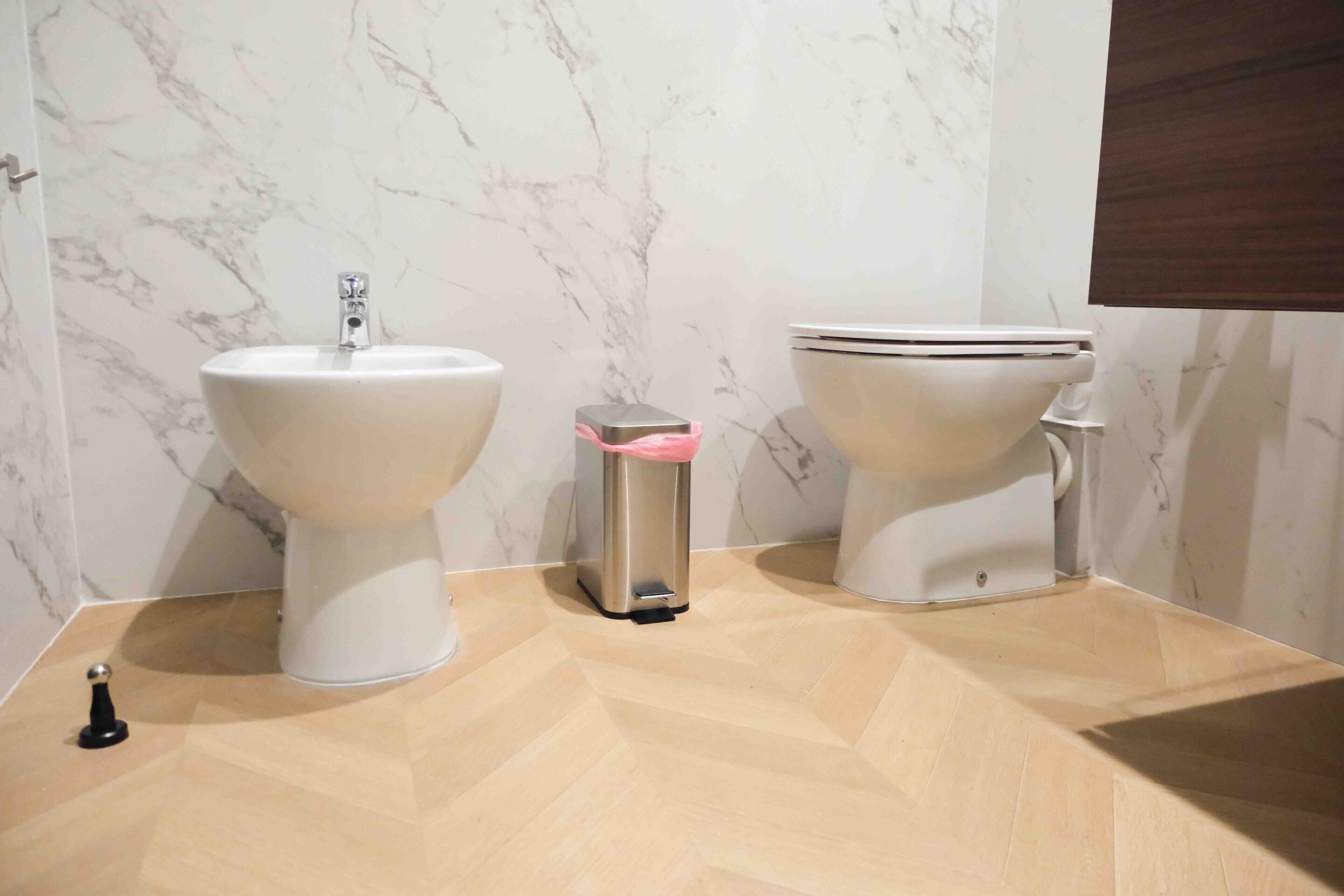 A bathroom with a marble wall, a bidet, a toilet, and a trash bin.