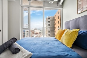 Quarto com uma grande janela, cama com roupa de cama azul e vista para edifícios lá fora.