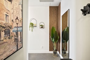 Um corredor com um quadro grande emoldurado, dois espelhos fixados na parede e dois vasos de plantas.