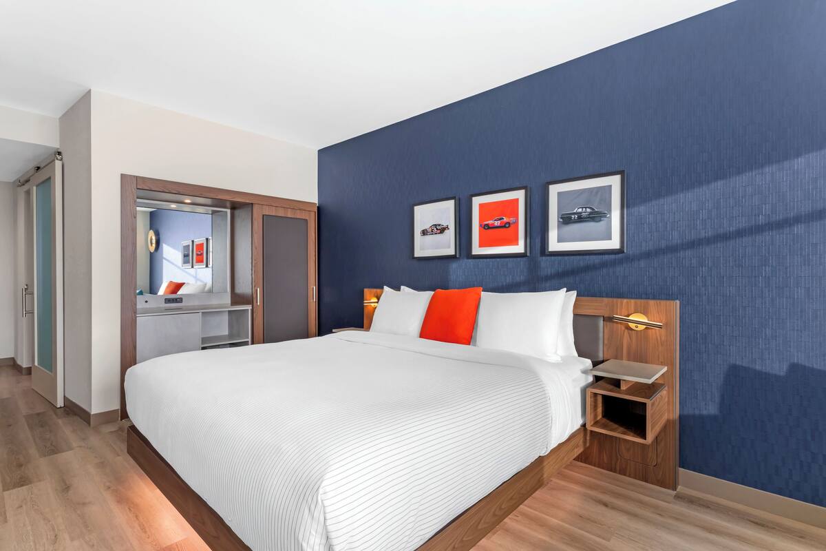Ein Hotelzimmer mit einem großen Bett, einem Nachttisch aus Holz und einem Schreibtisch mit Stuhl. An der Wand hängen drei gerahmte Bilder.