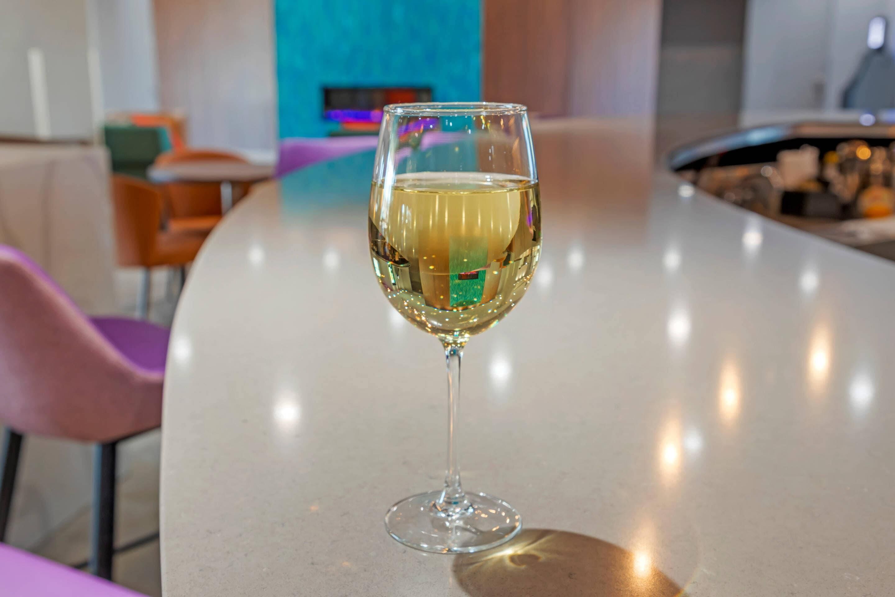 Una copa de vino blanco sobre una barra con sillas y una pared colorida al fondo.