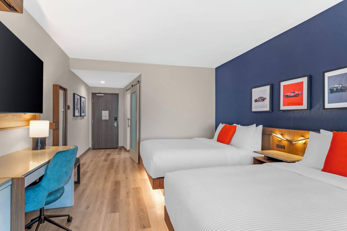 Ein Hotelzimmer mit zwei Betten, einem Schreibtisch mit Stuhl, einem Fernseher und gerahmten Bildern an der Wand.
