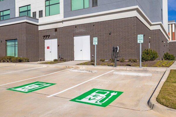 A parking area with electric vehicle charging stations, marked parking spaces, and a building with a brick facade.