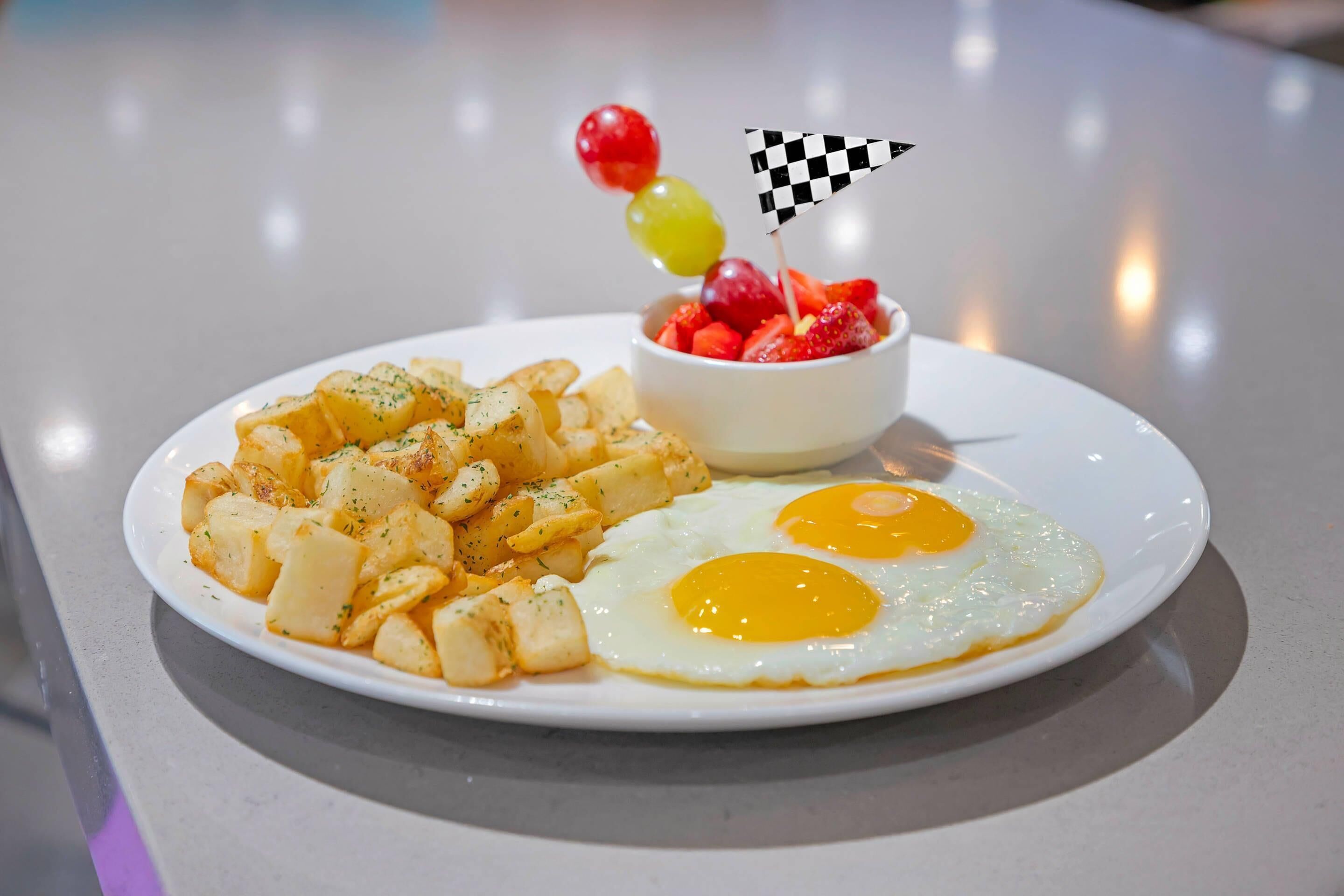 Ein Teller mit Spiegeleiern, Bratkartoffeln und einer kleinen Schale Obst.