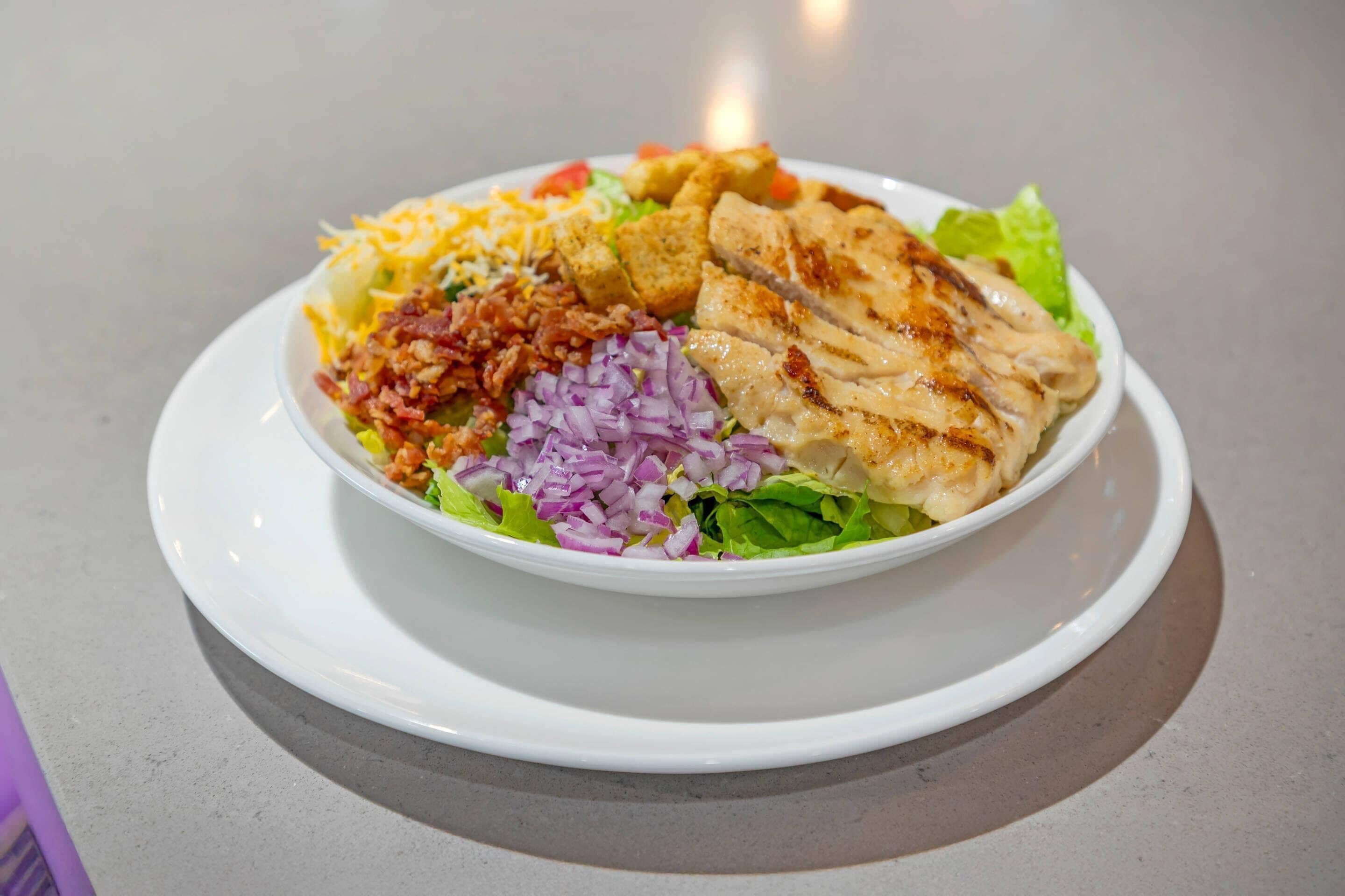 Un plato blanco con ensalada de lechuga, cebollas rojas, queso rallado, crutones y pollo a la parrilla.