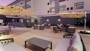 An outdoor seating area with tables, chairs, and umbrellas, surrounded by a building with multiple windows. - Cambria Hotel Northlake Conference Center - Roanoke (Northlake)