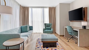 A modern hotel room with a sofa, ottoman, desk, chair, and a flat-screen TV. - Cambria Hotel Northlake Conference Center - Roanoke (Northlake)