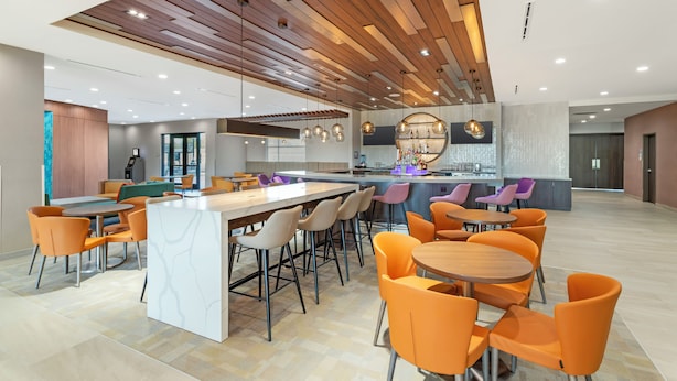 A modern kitchen with a central island, bar seating, and orange chairs. - Cambria Hotel Northlake Conference Center - Roanoke (Northlake)