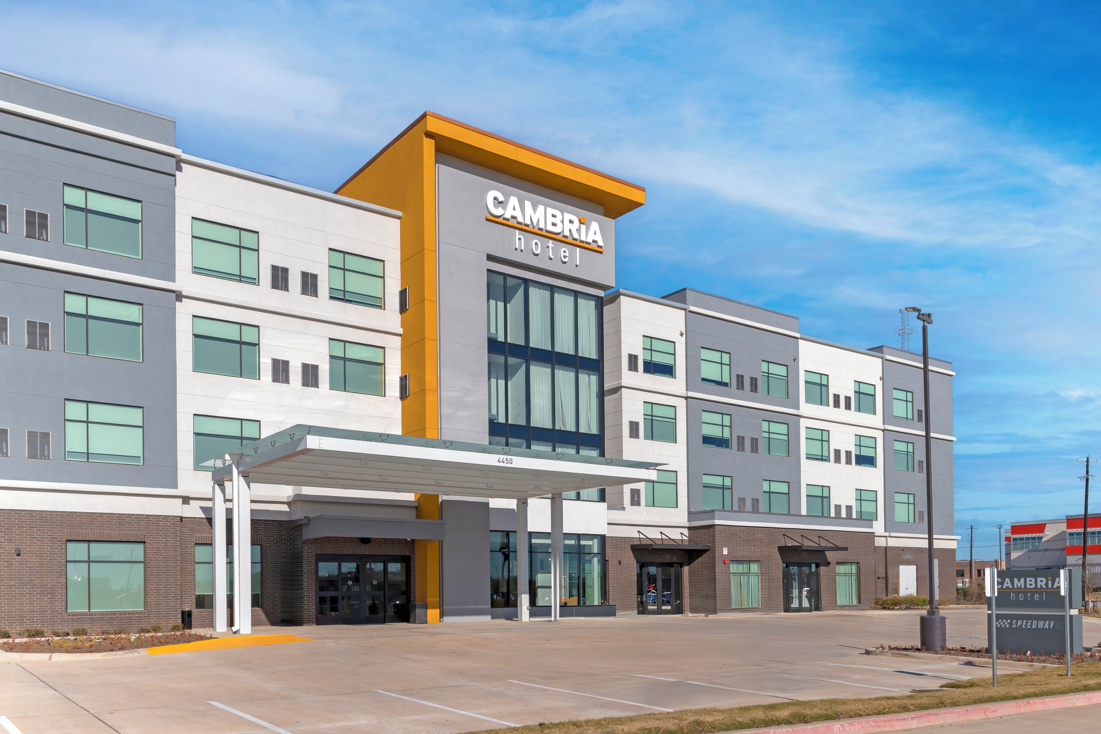 A modern hotel with a prominent "CAMBRIA hotel" sign, a covered entrance, and a parking area.