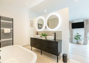 Une salle de bain moderne avec une baignoire sur pieds, un meuble-lavabo noir doté de deux vasques et un grand miroir avec un éclairage circulaire.