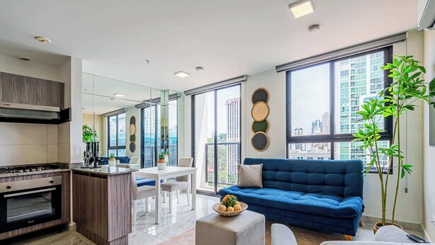 A modern kitchen with a gas stove, a dining area with a white table and chairs, a blue sofa, and large windows offering city views. - w* | Sophisticated 1BR on Calle 50 w/ View (Panamá)