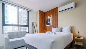 A modern hotel room with a large bed, a grey sofa, a wooden accent wall, and a city view through the window. - w* | Sophisticated 1BR on Calle 50 w/ View (Panamá)