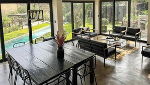 Een moderne woonkamer met een eettafel, zwarte leren banken en grote schuifpuien naar een zwembad en tuin.