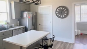 A modern kitchen with a central island, bar stools, a hanging light fixture, a refrigerator, and a wall clock. - Charming 2-bedroom farmhouse in delightful Auburn with AC, WiFi (Auburn)