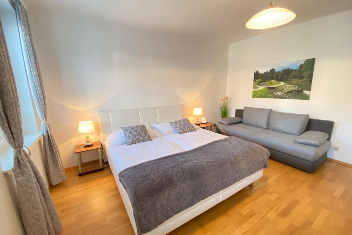 A bedroom with a bed, a grey sofa, a nightstand with a lamp, and a wall-mounted picture.