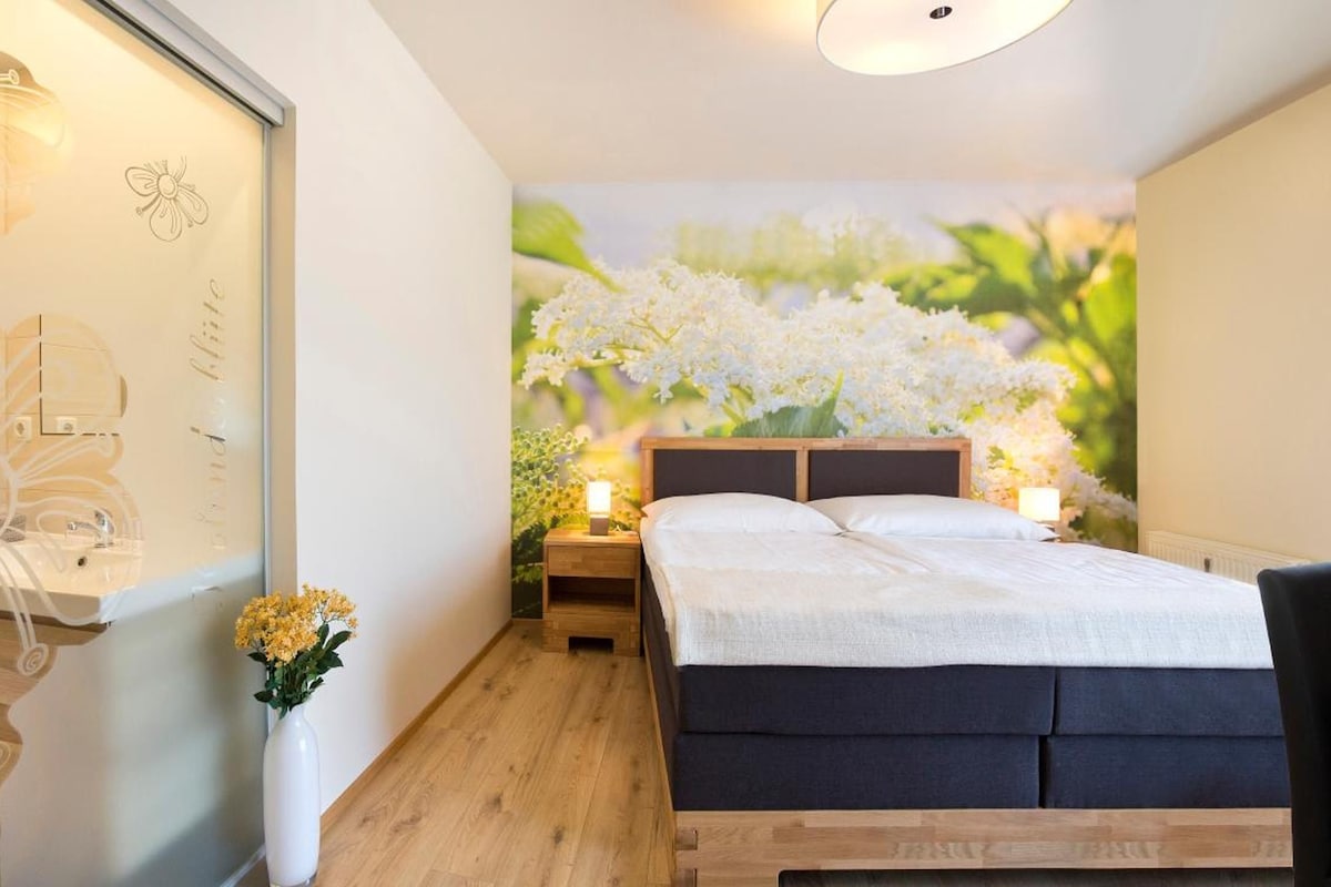 A bedroom with a bed, bedside tables, a nightstand, a vase with flowers, and a wall with a floral mural.