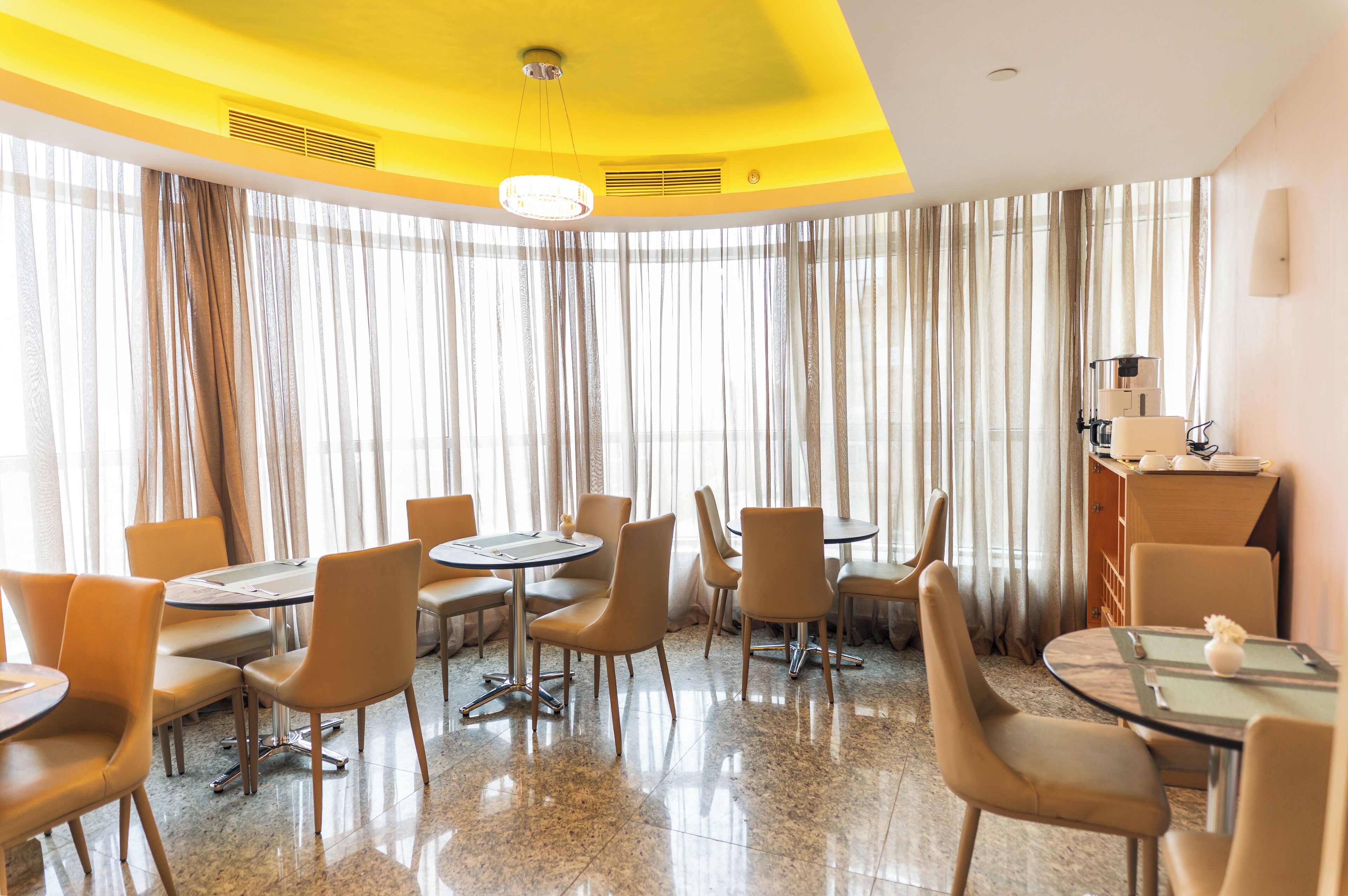 A hotel dining area with round tables and chairs, a central chandelier, and a large window with curtains.