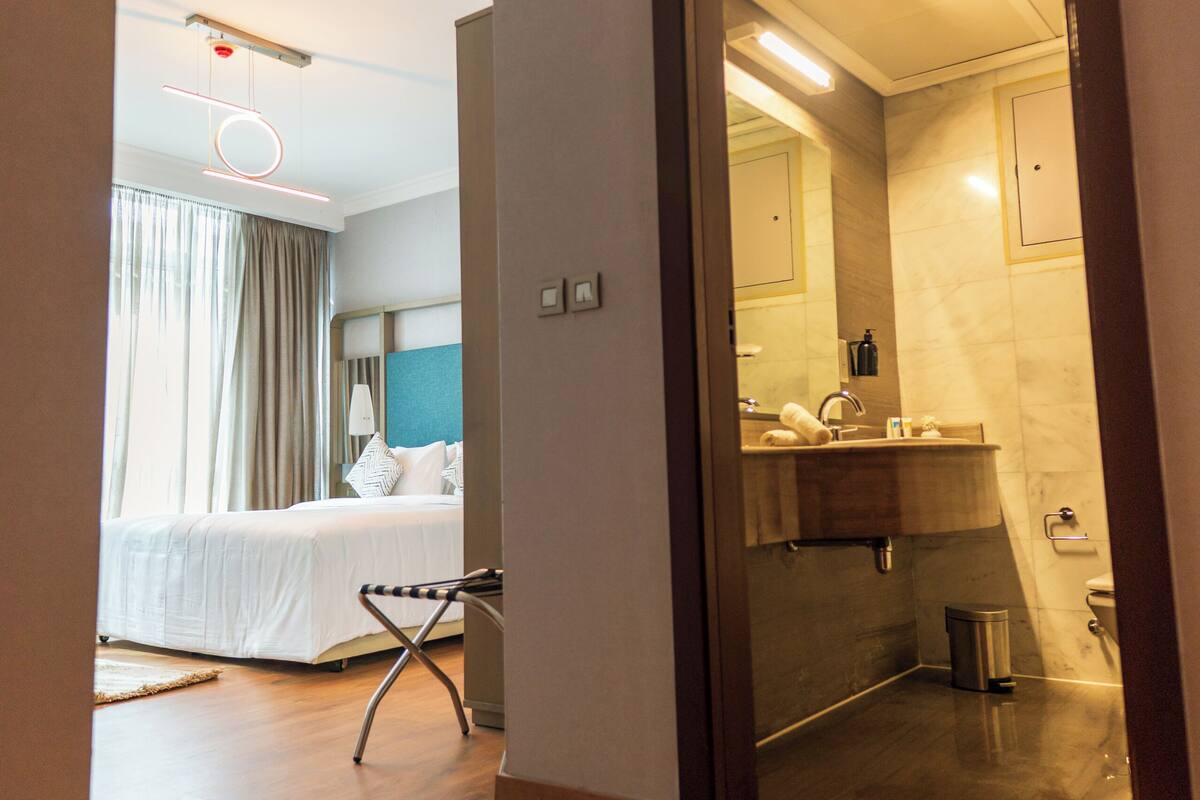 A hotel room with a bed, a bedside table, a mirror, a bathroom with a sink and toilet, and a wooden floor.