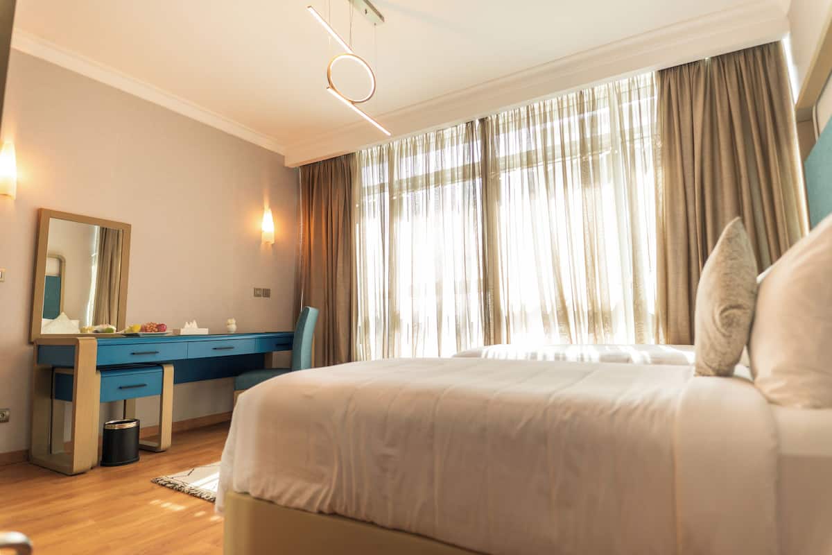 A hotel room with a large bed, a desk with a chair, a mirror, and a window with curtains.