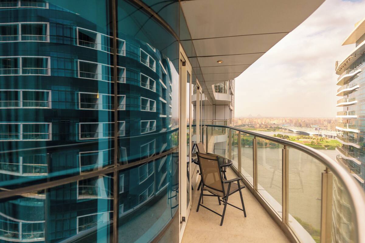A balcony with a glass railing, a chair, and a view of a modern building and a river.
