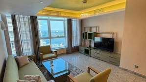 Una sala de estar moderna con sofá, mesa de centro y sillones, televisor de pantalla plana y vistas a la ciudad a través de amplias ventanas.