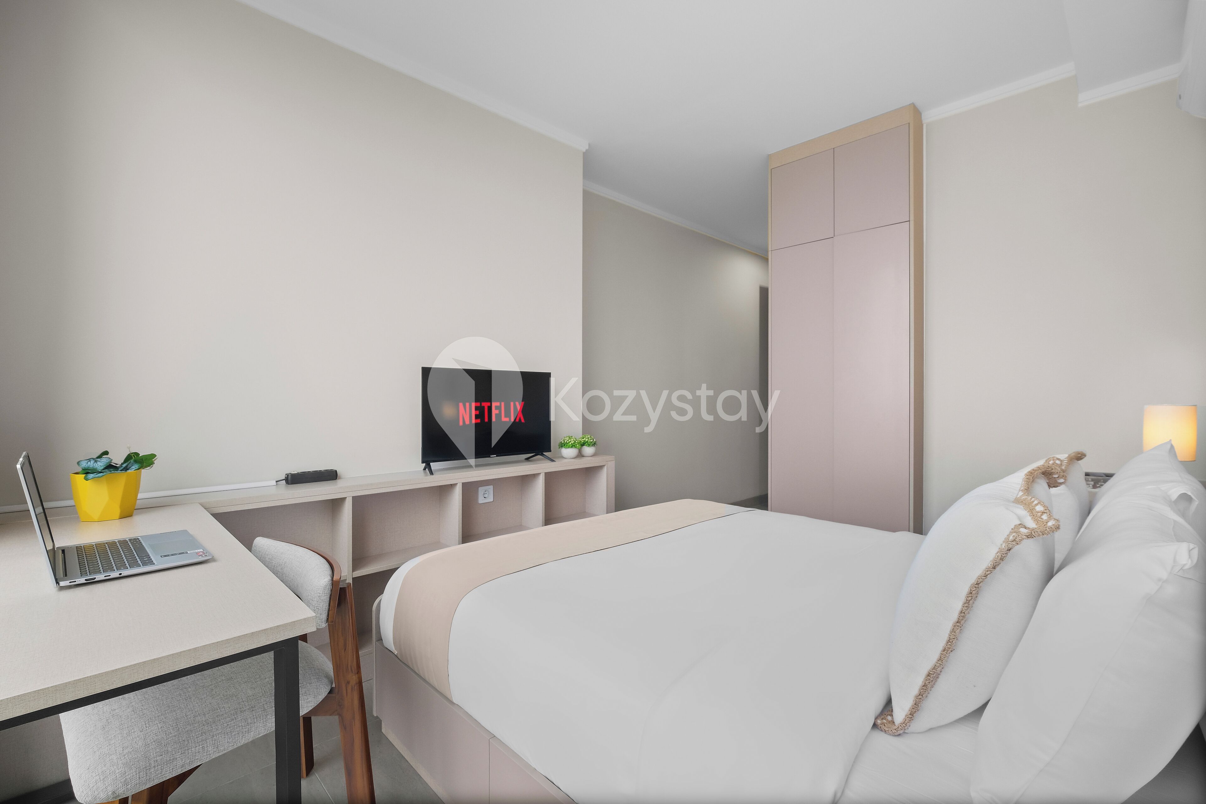 A modern hotel room with a bed, a desk with a laptop, a TV, and a small plant.