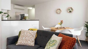 Una sala de estar moderna con un sofá gris, una mesa de comedor con sillas blancas y una cocina al fondo.
