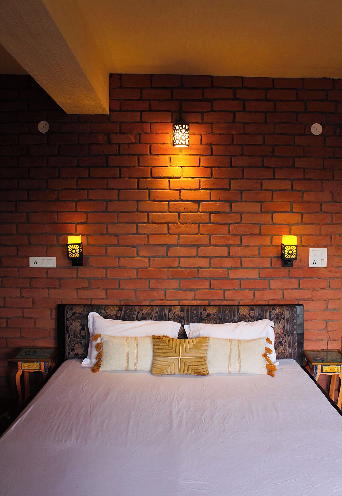 A room with a brick wall, a bed with white bedding, two wall-mounted lamps, and a wooden headboard.