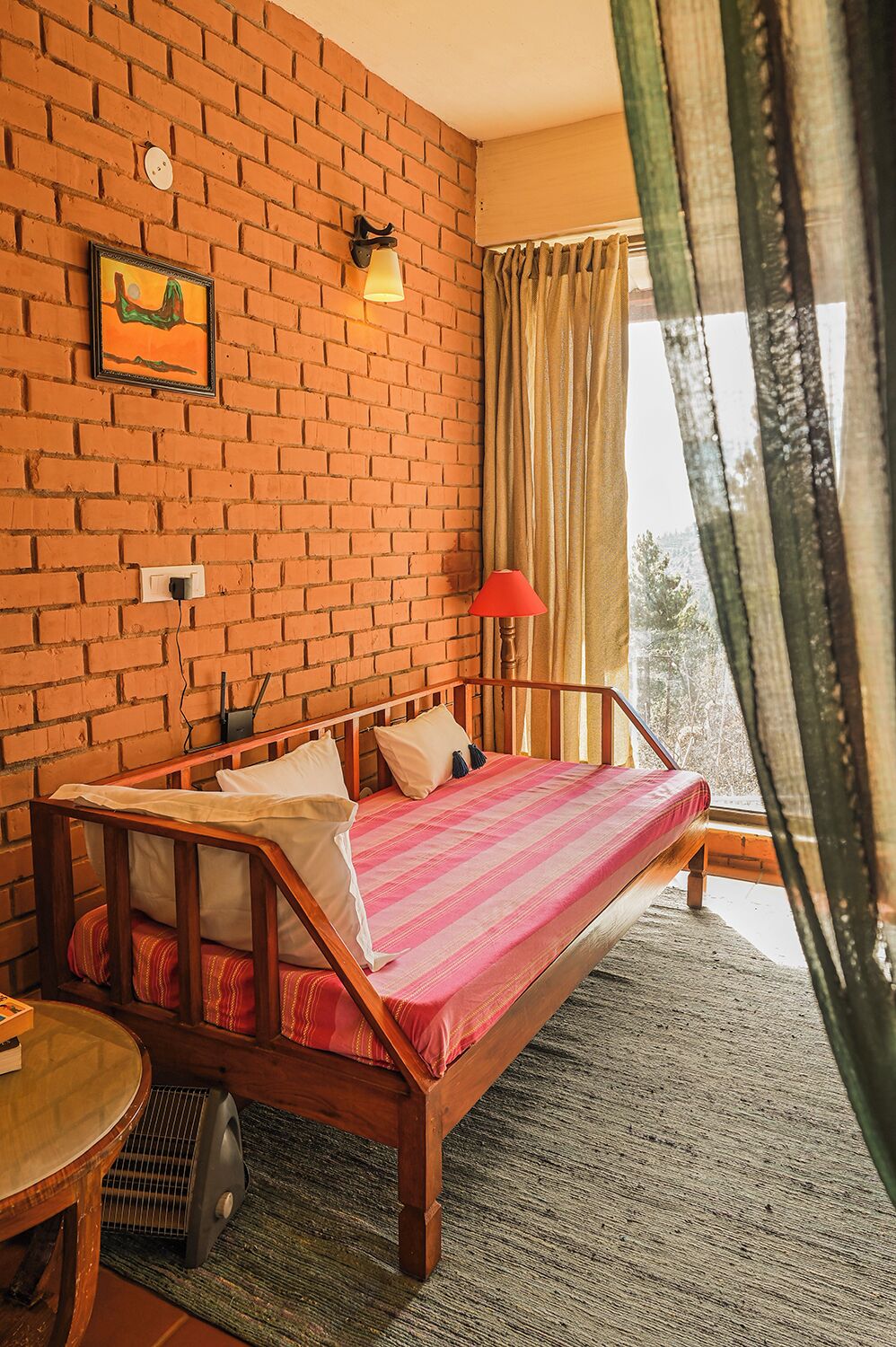 Une pièce avec un mur en briques, un lit recouvert d’une couverture à rayures roses et blanches, une table de chevet en bois, une lampe rouge et une fenêtre avec des rideaux.