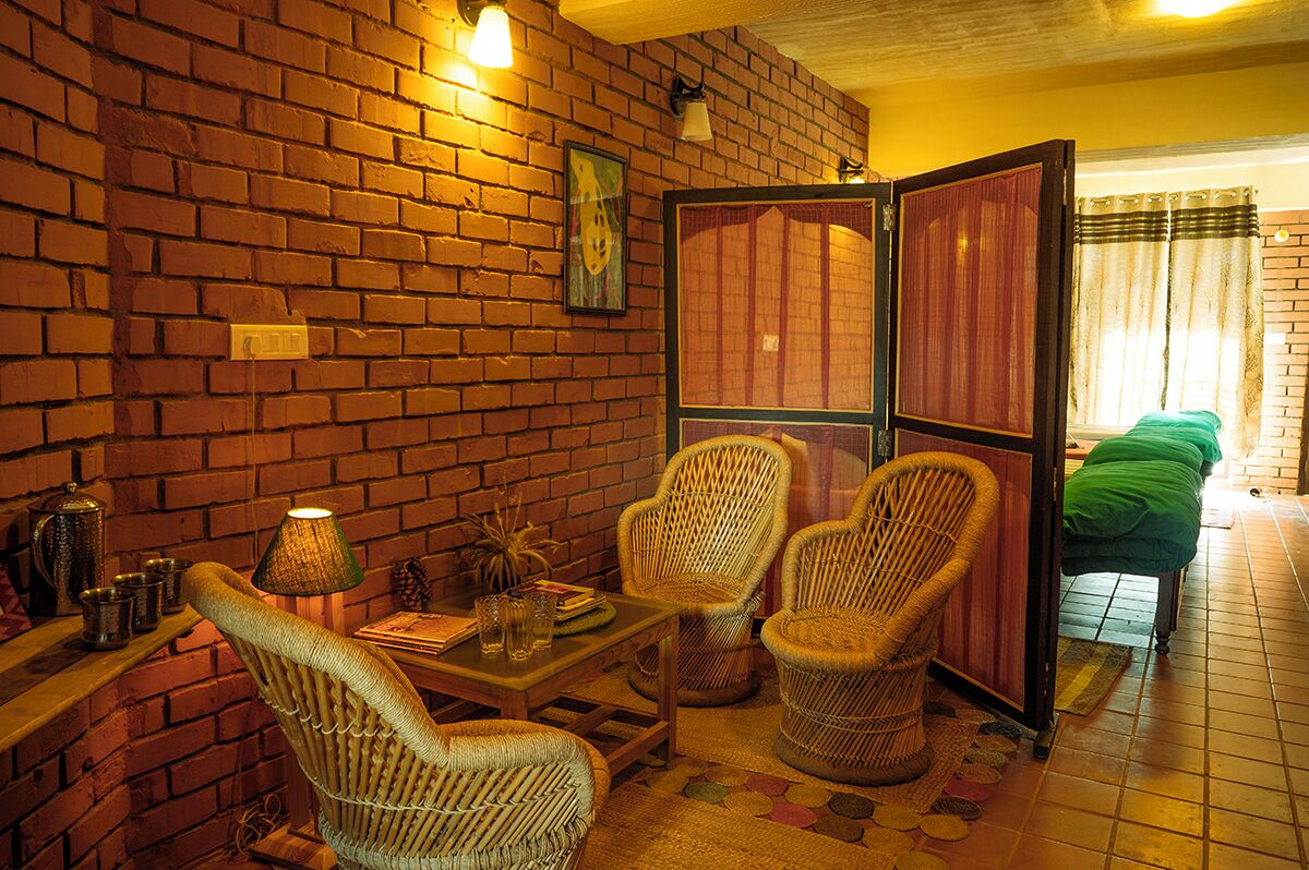A room with a brick wall, wicker chairs, a table with drinks, and a bed with a green blanket.
