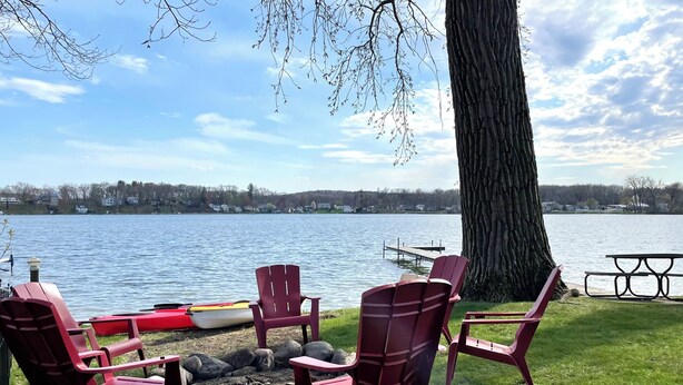 Et opholdsområde ved søen med Adirondack-stole og en kano, omgivet af græsplæne og et stort træ.