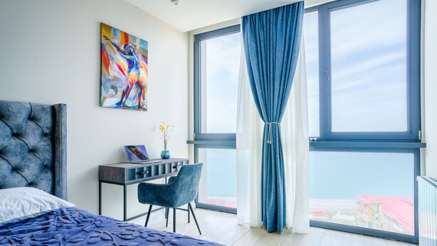 Un dormitorio con una cama, un escritorio y una silla. Hay un ventanal con cortinas y vistas a edificios.
