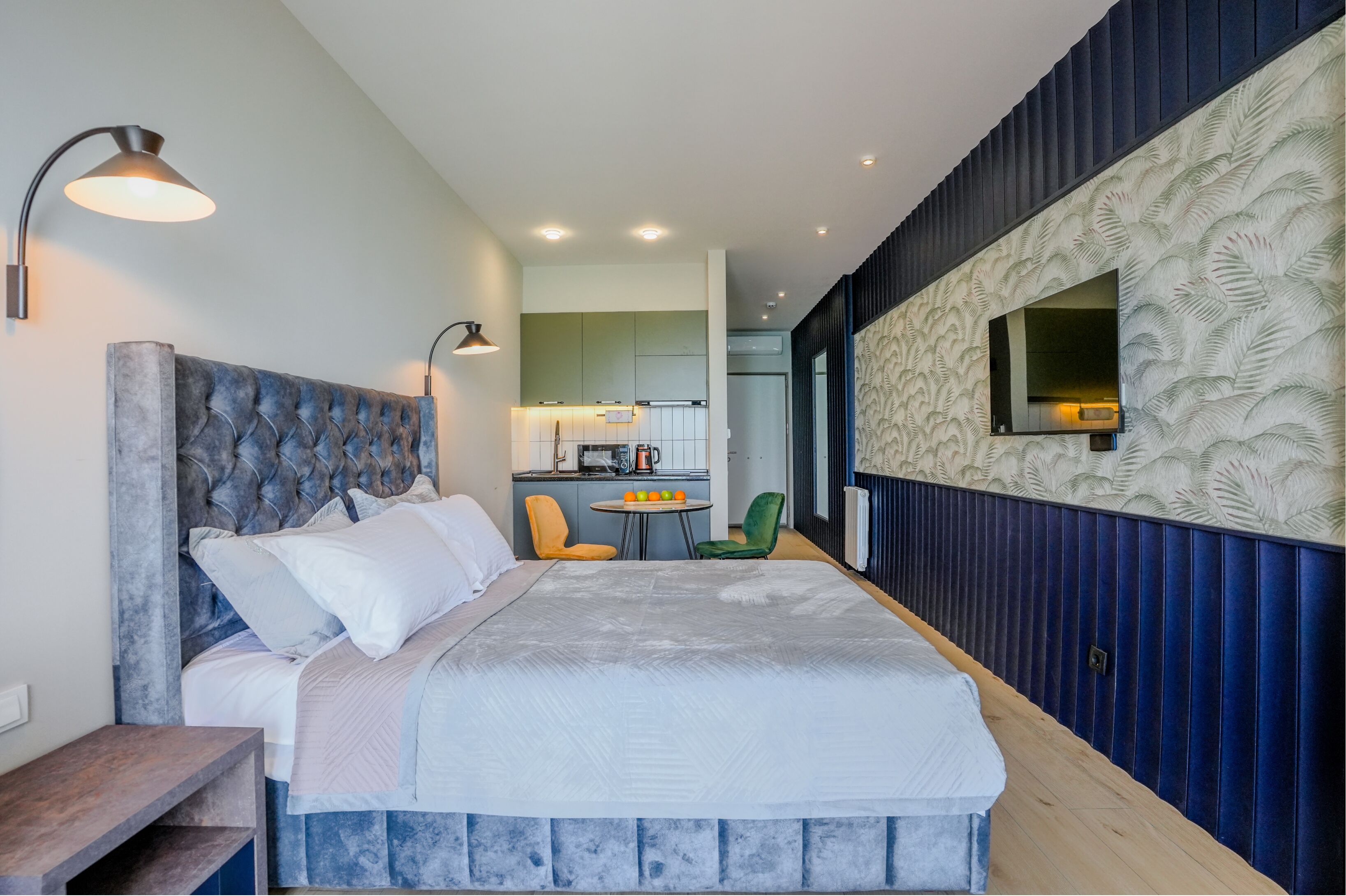 A bedroom with a large bed, a small table, and a chair, a kitchenette in the background, and a patterned wall.
