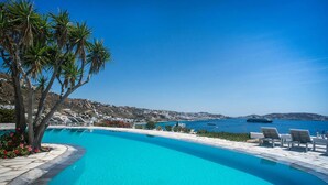 A clear blue pool with a curved edge, surrounded by palm trees and lounge chairs, overlooking a coastal landscape with buildings and boats. - My Rental Homes - Viila Rose 2  only 1 km from Agios Stefanos beach. Shared pool (Mykonos)