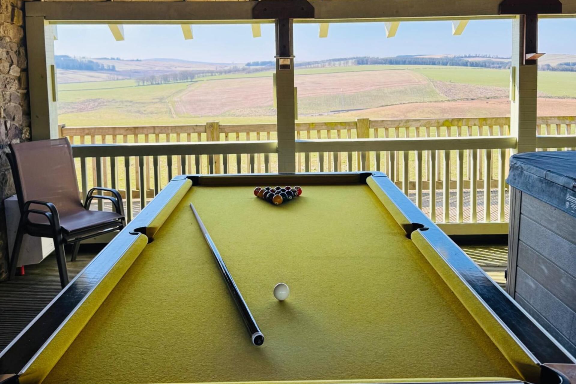 A pool table with a ball, a chair, and a view of a rural landscape through a wooden railing.