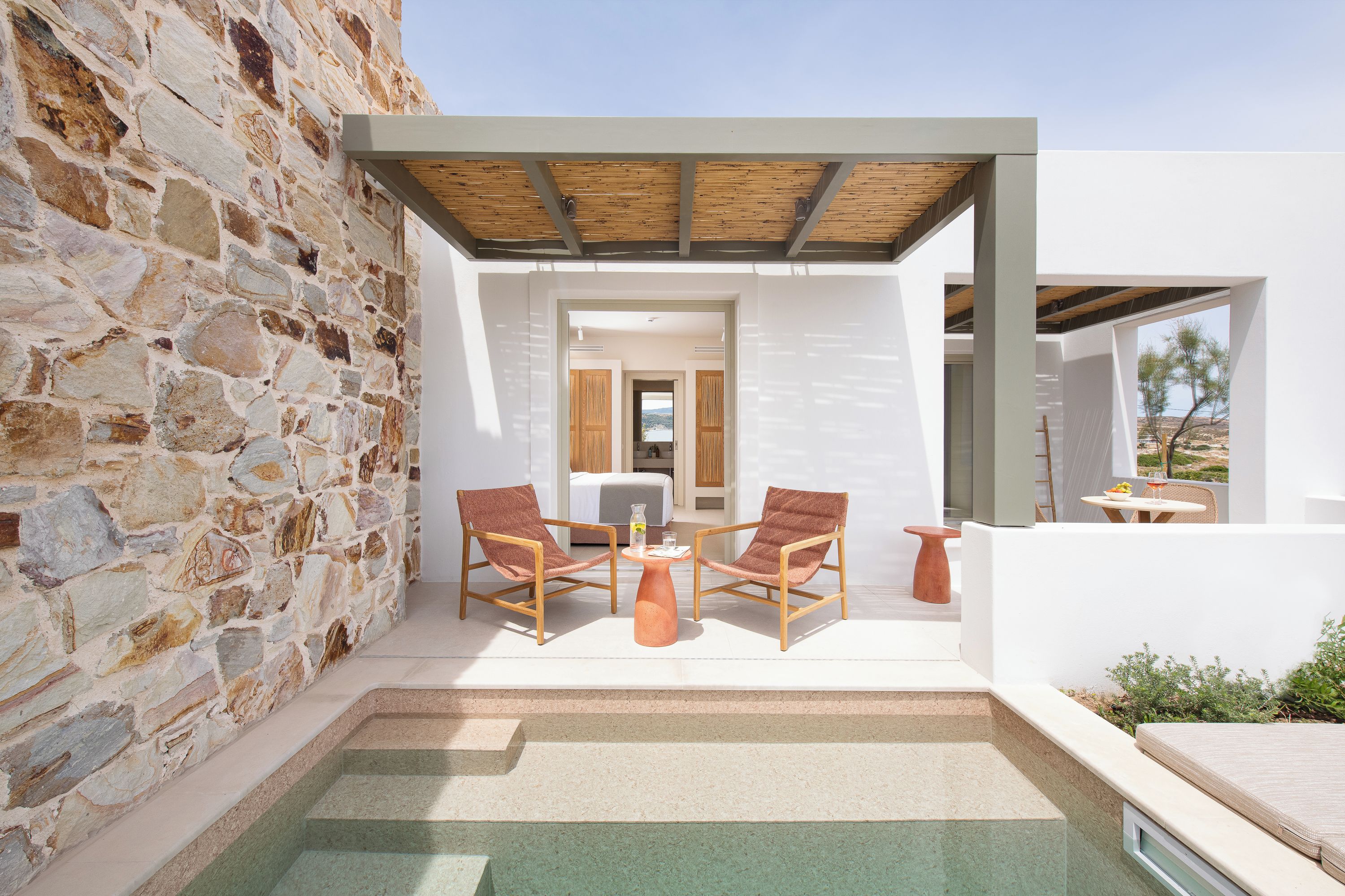 A modern outdoor seating area with stone walls, a wooden pergola, and a view of the interior.