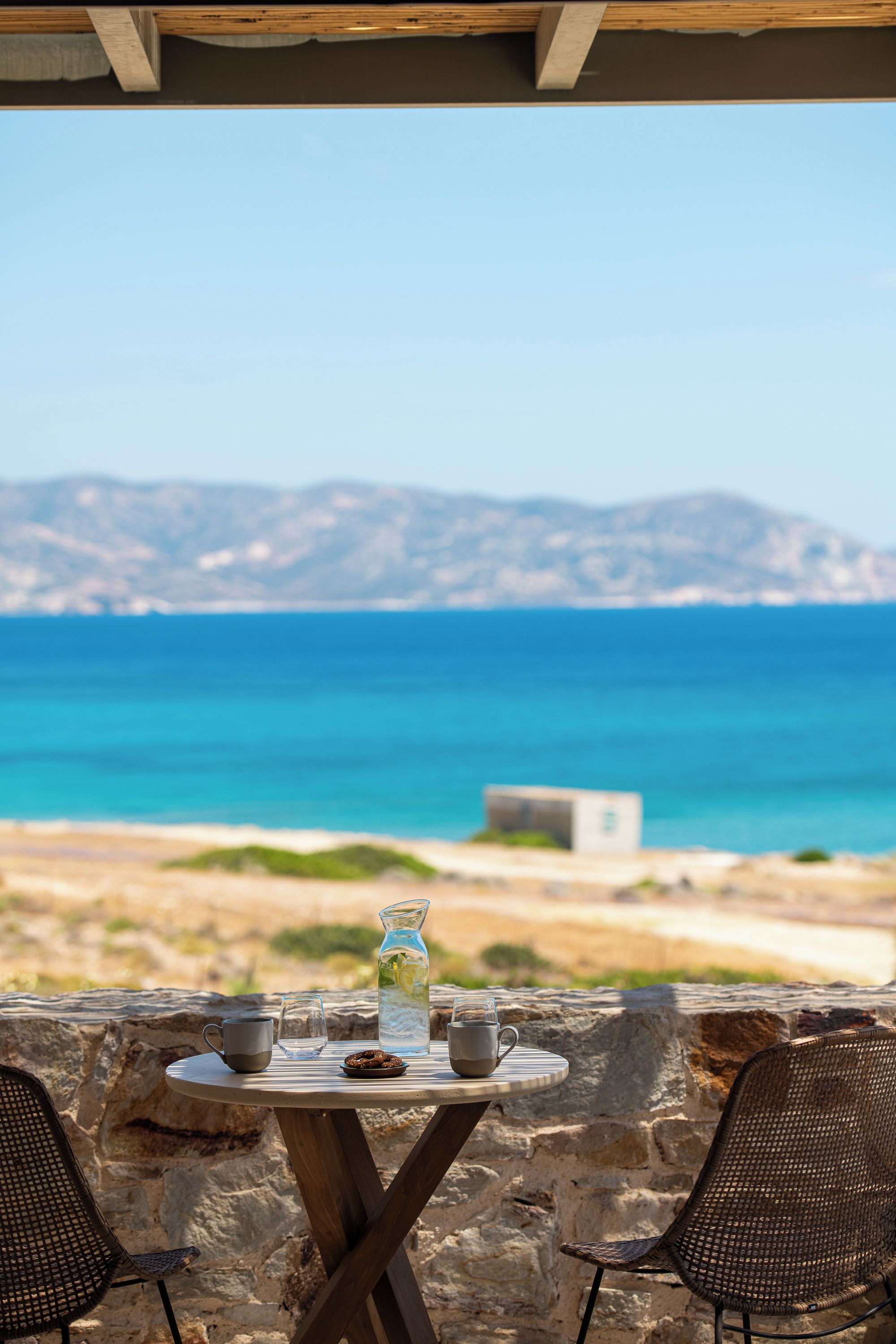 A table with a glass pitcher, two cups, and a plate of cookies, overlooking a coastal view with a clear sky and distant mountains.