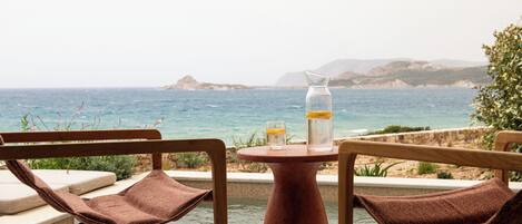 Zona junto a la piscina con dos sillas de madera y una mesa, con vistas a un cuerpo de agua y tierra distante.