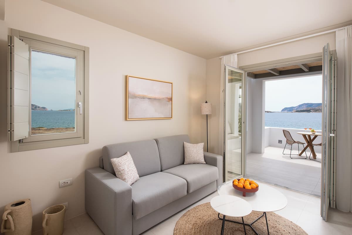 A modern living room with a grey sofa, a small table with oranges, and a view of the sea through an open door.