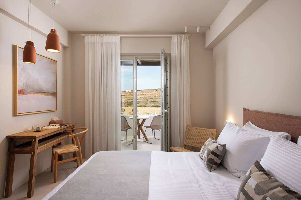 A bedroom with a bed, a desk, and a chair. There is a view of a beach through the window.