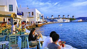 A seaside restaurant with outdoor seating overlooking the water, featuring white buildings and windmills in the background.